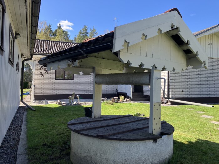 Vit, dekorativ överbyggnad för brunn med takkonstruktion och vev, belägen på en gräsmatta mellan två hus.