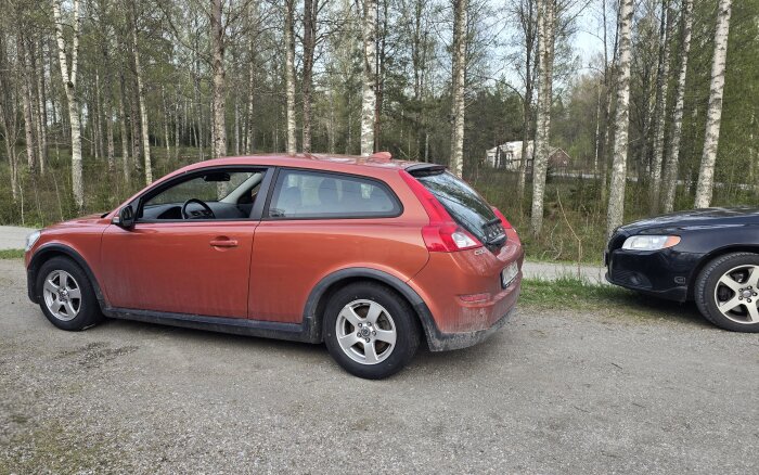 Röd bil med nya sommarhjul och ballongdäck parkerad på grusväg, omgiven av björkträd, med en del av en annan bil synlig.