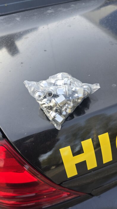 Plastförpackning med flera nya silverfärgade muttrar ligger ovanpå en mörk bilbaklucka med texten "HI" i gult nära bakljusen.