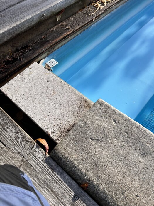 Poolkant med skadade plattor vid vattenytan samt närliggande trädäck i trä.