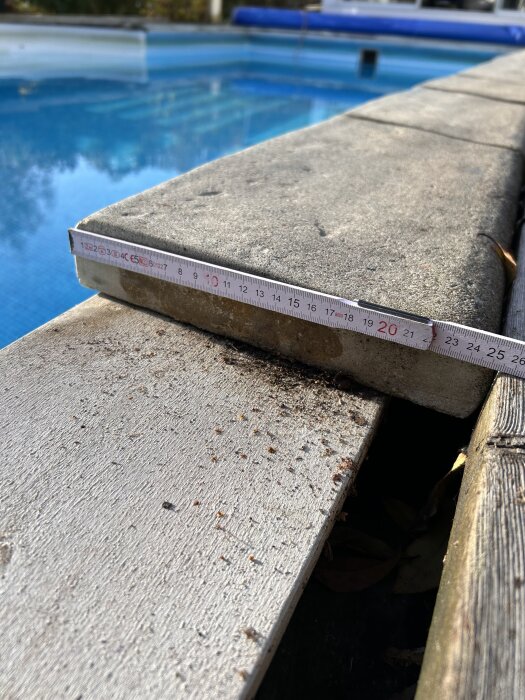 Måttband mäter plattans tjocklek vid en poolkant, intill tryckimpregnerade brädor, visar behov av utbyte på grund av dålig limning och utseende.