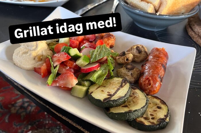Grillad chorizo med sallad, hummus, grillade champinjoner och zucchini på tallrik, bröd i bakgrunden. Text: "Grillat idag med!".