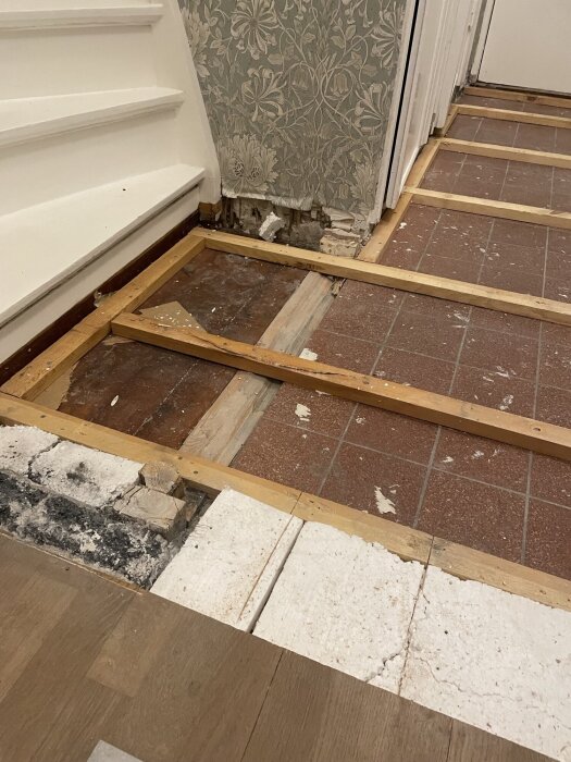 Trägolv och gammalt klinkergolv vid trappan, med träreglar som blottats i renoveringsprocessen. Tapetserad vägg i bakgrunden.