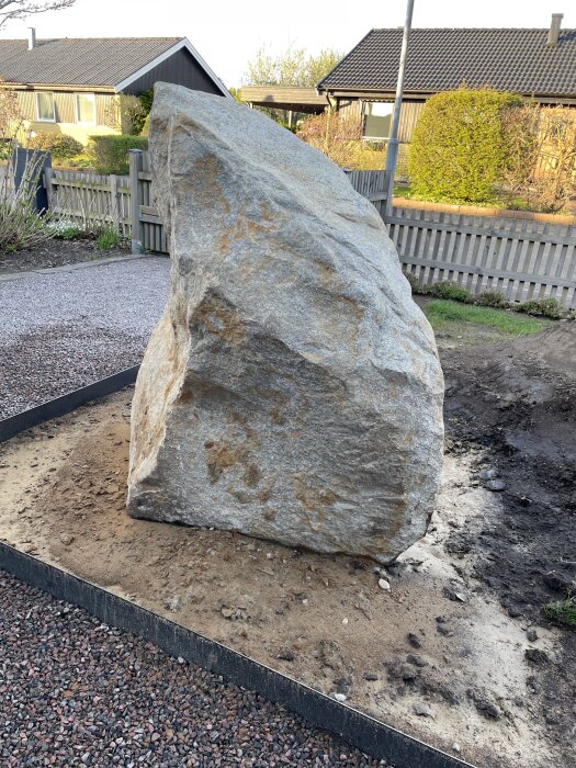 Stor sten placerad på en sandbädd omgiven av grus, i en trädgårdsmiljö med staket och hus i bakgrunden.