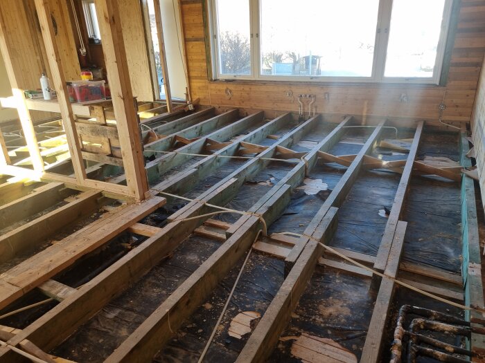 Golvbjälkar synliga i ett rum under renovering, med borttagen kutterspån och påbörjat arbete med bjälklag och fönster.