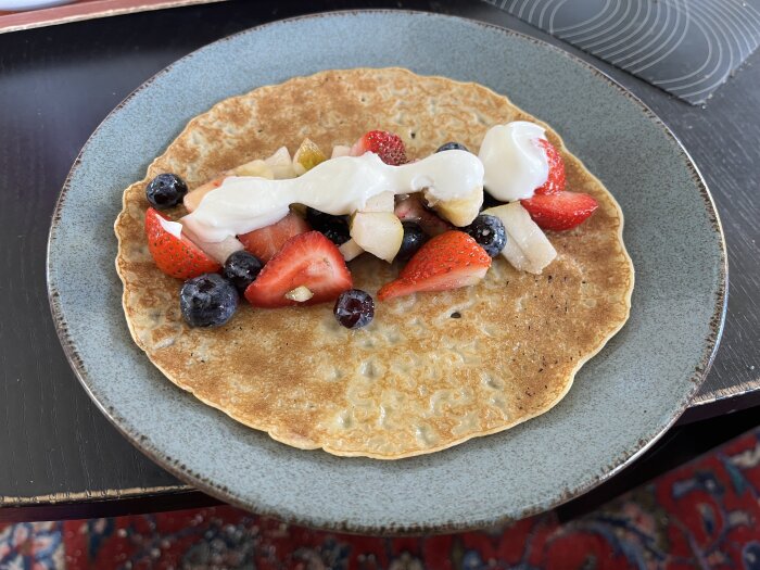 Tunnpannkaka med fruktsallad av päron, banan, blåbär och jordgubbar, toppad med vispad grädde, serverad på tallrik.