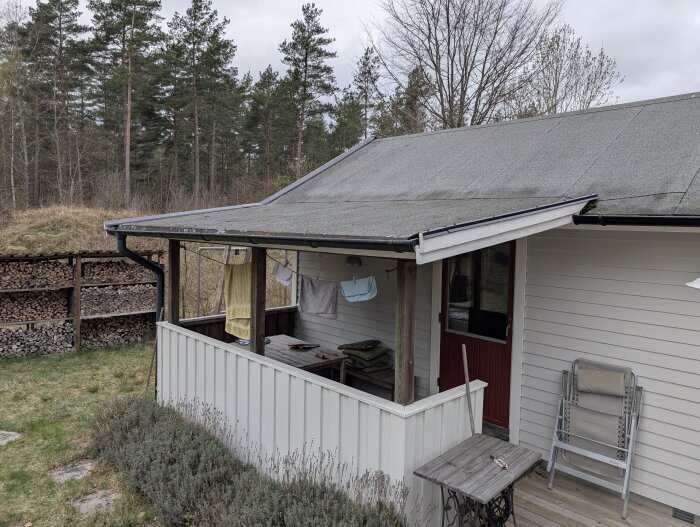 Uteplats med takpapp vid bostadshus, omgiven av lavendel och skogsbryn, med vedförråd i bakgrunden.