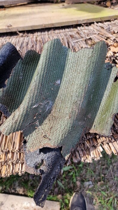 En bit av vågig takpapp med grönt ytskikt och svart undersida, liggande på ett underlag av stråmaterial.