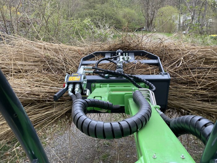 En grön maskin arbetar med att samla och kratta ihop vass på en grusväg omgiven av träd och buskar.