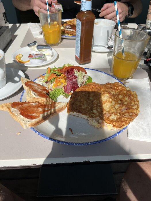Frukost med omelett, grönsallad och toast med sylt på en tallrik. Apelsinjuice och kaffekopp på bordet.