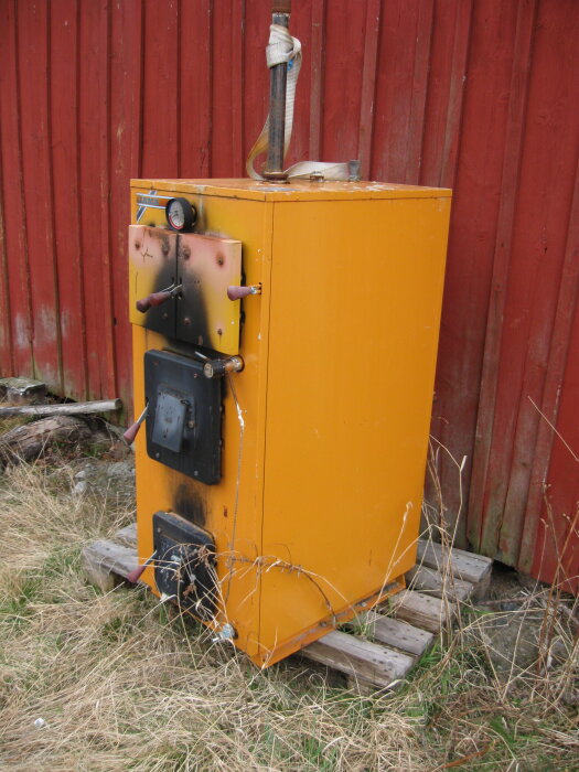 Orange vedpanna från 1992 med 25kW på olja och 40kW på ved, placerad utomhus mot röd vägg på träpallar.