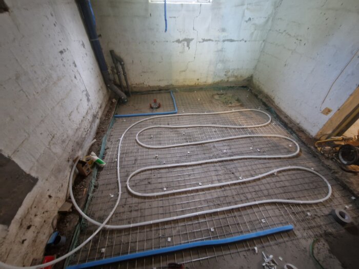Golvvärmeslingor installerade på ett nät på ett betonggolv i en källare, med synliga rör längs väggarna och diverse byggverktyg runtomkring.