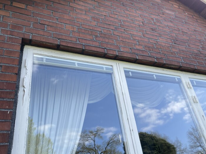 Tegelbalk över fönsterparti, vitmålade fönsterkarmar, himmel och träd reflekteras i glaset.