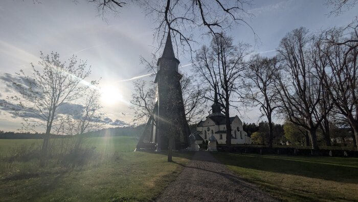 Kyrka vid en grusväg omgiven av höga träd en solig dag, med solen lågt på himlen och ängar i bakgrunden.
