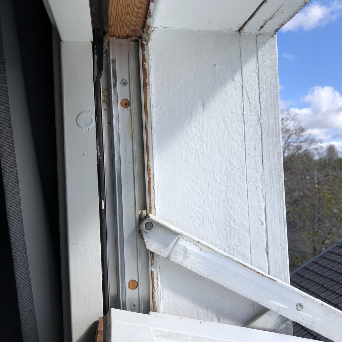 Ett trasigt fönster med en avbruten vit metallarm mot träkarm och blå himmel i bakgrunden.