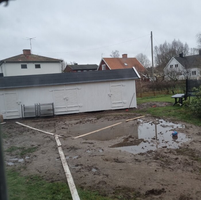 Lerig tomt med pölar framför vit byggnad, omgiven av villaområde. Brädor korsar marken; gräsområde närmast kameran. En boll ligger nära vattenpölen.