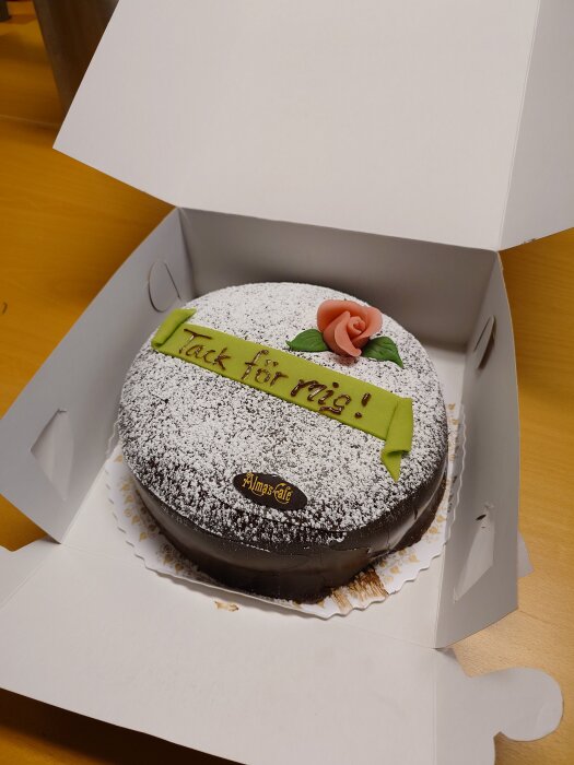 Chokladtårta med grön marsipanband dekorerad med texten "Tack för mig!" och en ros, i en öppen vit kartong.