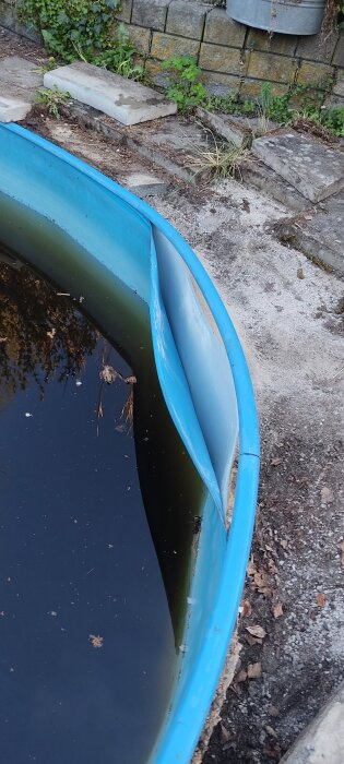 Skadad poolkant med blå liner som har lossnat och sjunkit. Vattenytan är delvis täckt av löv och omgiven av stenplattor och växtlighet.
