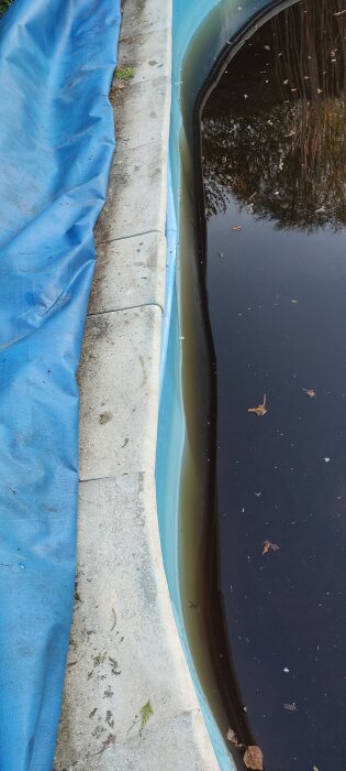 Skadad poolkant med plastliner och mörkt vatten, delvis täckt av en blå presenning, visar tecken på översvämning och skevhet.