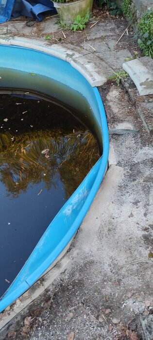 Skadad pool med blå liner och smutsig vattenyta, omgiven av betong och växtlighet. Kanten är ojämn och marken runtom är exponerad.