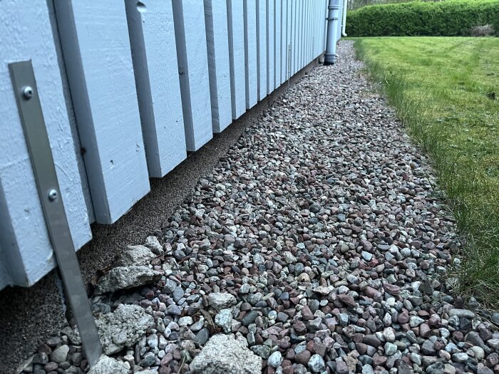 Metallband längs husets grund med små betongkockor vid kanten, omgiven av grus. Fråga om funktion och koppling till husets sjunkproblem.