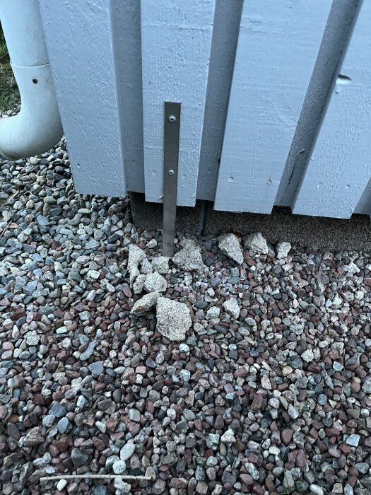 Metallband fastsatt på en husvägg med synliga betongklumpar och grus på marken.