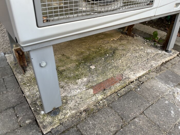 Luftvärmepump placerad på en sliten och ojämn betonggrund, omgiven av plattor.