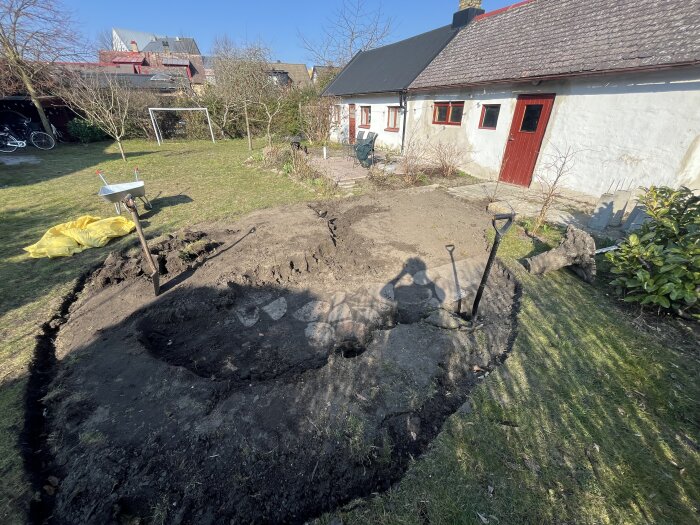 Trädgårdsrenovering med uppgrävd mark, spadar och skottkärra, intill ett hus med röd dörr och fönster.