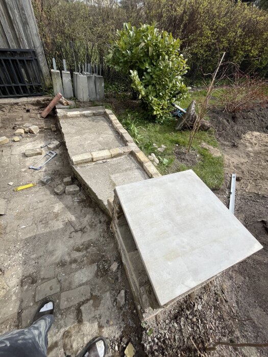 Trädgårdsrenovering med nylagd grund av betongplattor och Lomma tegel, omgiven av gröna buskar och växter. Verktyg och material synliga på marken.