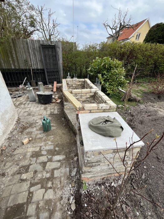 Stenlagd uteplats under renovering med tegel och murade väggar, omgiven av buskar och ett gult hus i bakgrunden.