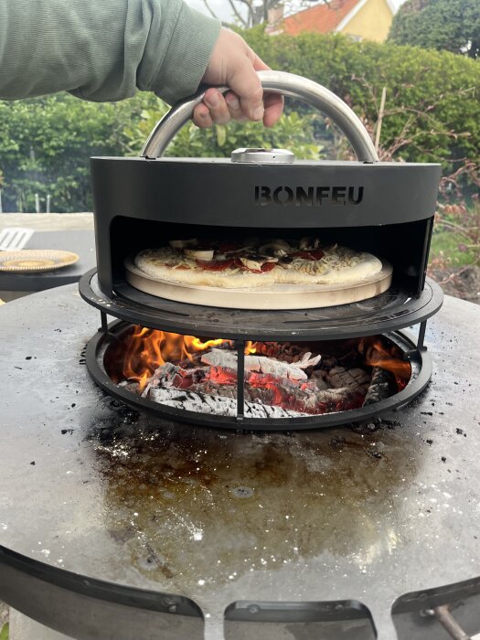 Utekök med eldstad och pizzaugn där en person lagar pizza; grönska och hus syns i bakgrunden.