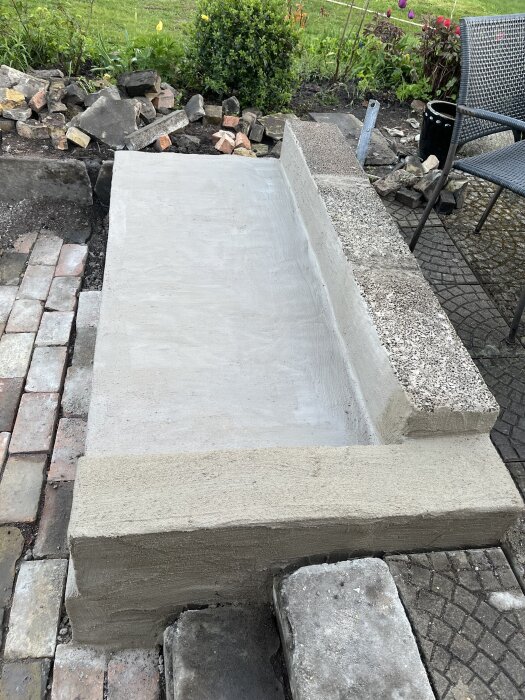 Betongplattform med omgivande stenläggning i en trädgård under renovering, omgiven av olika stenar och växter.