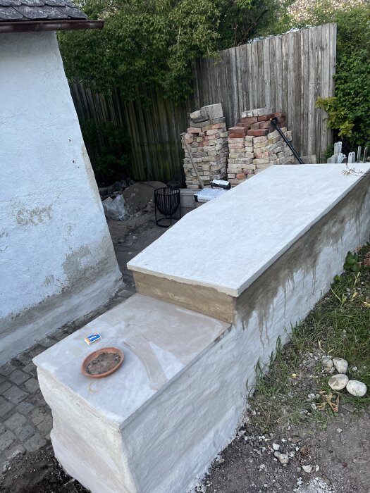 Halvmur i trädgård under renovering, omgiven av tegelsten, mattallrik, tändsticksask och grönt staket i bakgrunden.