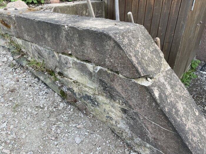 Gammal mur av betongblock från 70-talet med sprickor, delvis omgiven av grus och växtlighet, nära en träbyggnad.