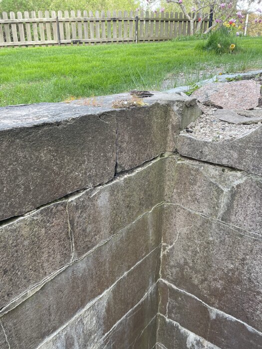 Gammal stenmur från 70-talet av hårda betongblock, delvis täckt av mossa, med trästaket och trädgård i bakgrunden.