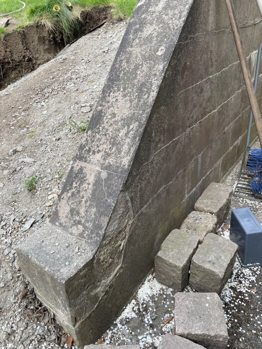 Gammal mur från 70-talet av hårda betongblock med sprickor, omgiven av grus och gräs.