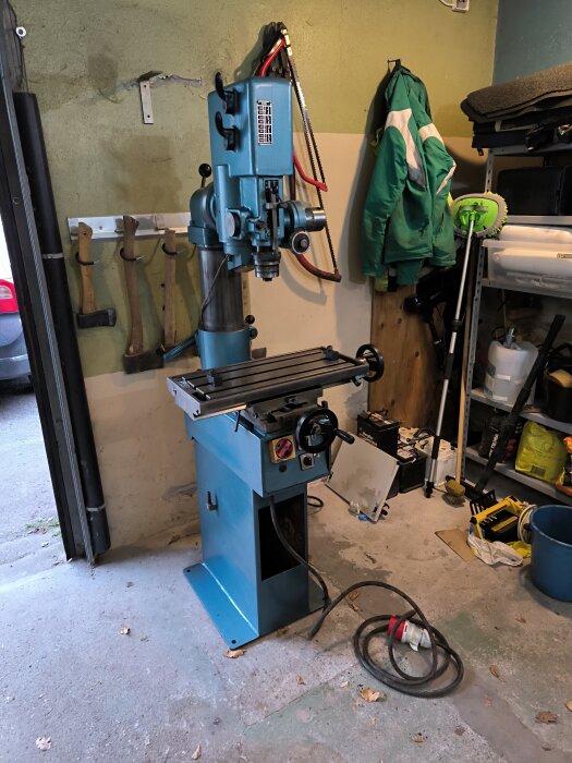Blå Arboga pelarborrmaskin i ett garage med verktyg och utrustning på väggarna, inklusive yxor och en såg. Kabel ligger på golvet framför maskinen.