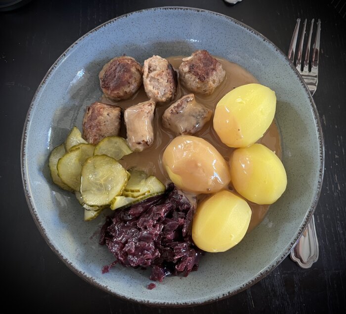 Köttbullar, kokt potatis, brunsås, rödkål och inlagd gurka på en tallrik.