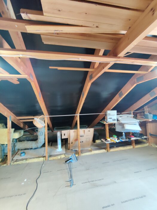 Kallvind med synliga takstolar, isolering, och olika byggmaterial syns. En kartong, en lampa och ventilationsrör ligger bland virket.