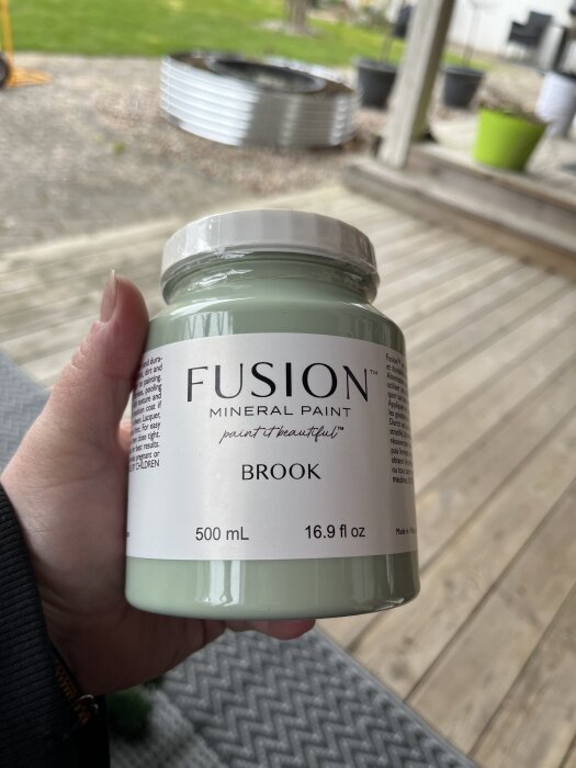 Burk med Fusion Mineral Paint i färgen Brook hålls i en hand, på en veranda.