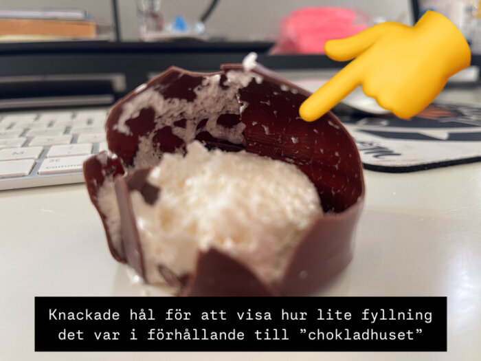En öppen chokladbit med lite fyllning syns, och en emojihand pekar på den. Bakgrunden visar ett tangentbord och suddig omgivning.