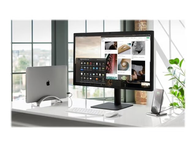 Kontorsbord med MacBook Air i hållare, stor skärmvisning, trådlöst tangentbord, mus, och iPhone i en docka, framför stort fönster.