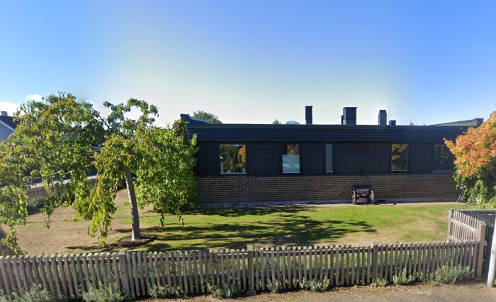 Framsida av hus med mörk fasad, ett staket framför och ett träd till vänster på tomten med gulgrönt gräs omkring.