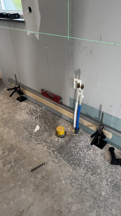 Vattenrör och monterade reglar på vägg under installation av bänkskåp i ett kök. Verktyg och klämmor används för justering.