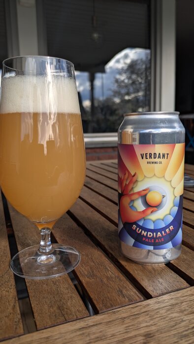Ett glas med ljus öl och en ölburk från Verdant Sundialer Pale Ale står på ett träbord. Båda med krämigt skum på ytan.