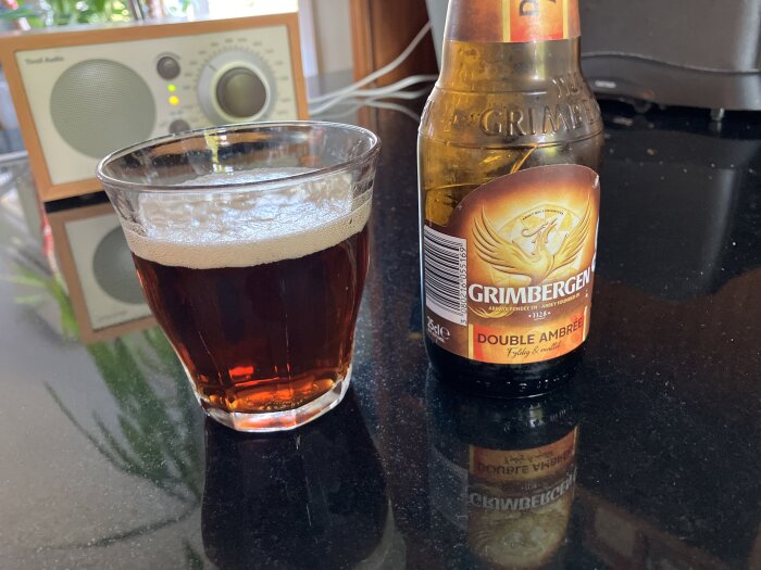 Glas med bärnstensfärgad öl bredvid en flaska Grimbergen Double Ambrée på ett svart bord, med en radio i bakgrunden.