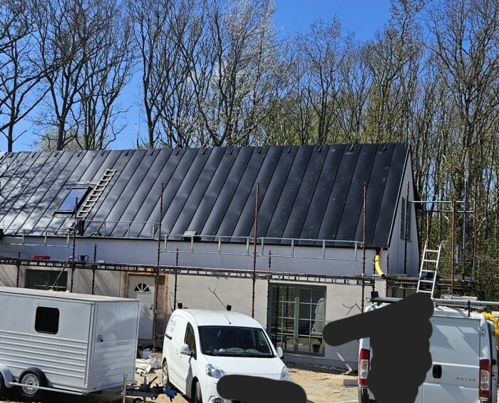 Tak med bucklor i plåtarna, byggnadsställningar runtom, ljus fasad och fordon framför huset under klarblå himmel.