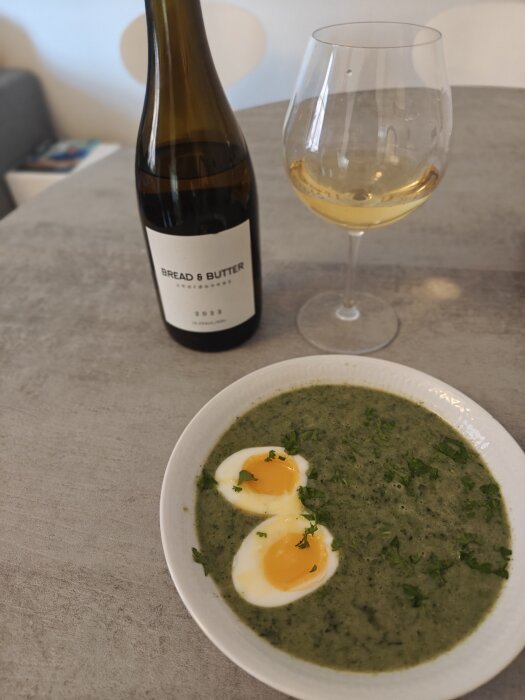 Sopptallrik med två halvor av kokt ägg, garnerad med örter, bredvid ett glas vitt vin och en vinflaska på ett bord.