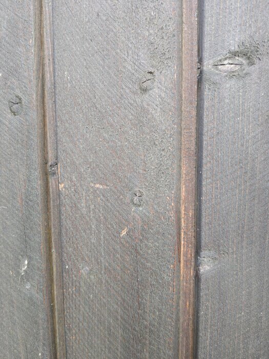 Närbild av träfasad med sliten, grå lasyr; ytan är ojämn och visar tecken på tidigare behandling och ålder.