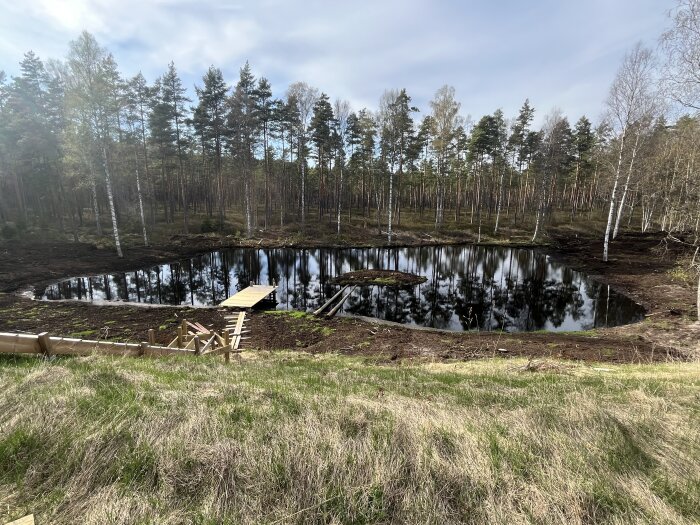 Damm i skogsmiljö med nybyggd trätrappa och brygga. Omgiven av träd och reflekterande vattenyta.